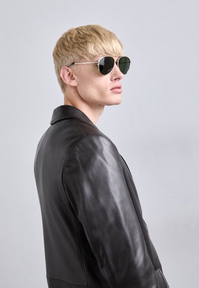 Blond person wearing dark aviator sunglasses and a black leather jacket, looking to the right against a plain light gray background.