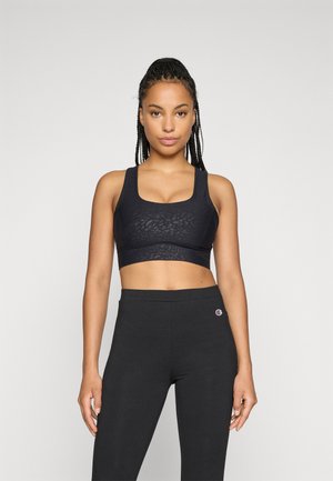 Kvinne med fletninger iført en svart mønstret sports-BH og svarte høyt midje leggings, som står mot en ensfarget bakgrunn.