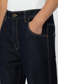 Dark blue denim jeans featuring a classic cut, stitched details in yellow, and front pocket with a subtle metal button closure.