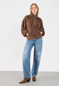 Brązowa, pluszowa bluza polarowa na zamek z wysokim kołnierzem i przednią kieszenią, zestawiona z jasnoniebieskimi, szerokimi spodniami dżinsowymi oraz brązowymi botkami na obcasie.