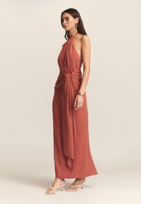 Hnědo-oranžové halter šaty s hlubokým výstřihem, uzlem v pase a volně splývajícími šaty až na zem. Modelka má na sobě béžové sandály.