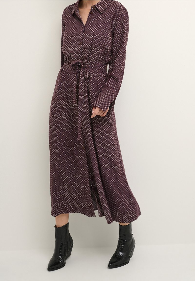 Długa, fioletowa sukienka z geometrycznym wzorem, zapięta na guziki z przodu, z pasekiem w talii. Zestawiona z czarnymi, szpiczastymi botkami za kostkę, wykonanymi z fakturowanej skóry.