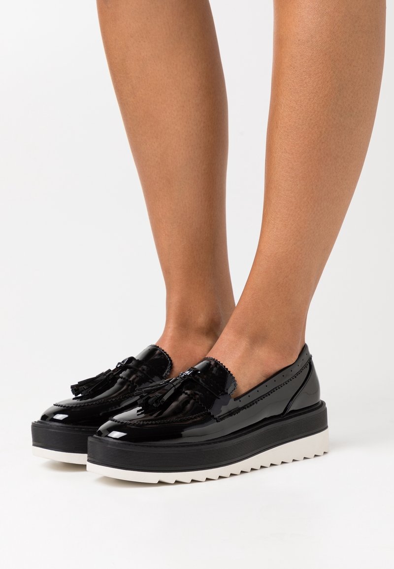 Jambes portant des mocassins à plateforme en cuir verni noir avec pampilles et semelles blanches épaisses et dentelées sur un fond blanc uni.
