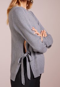 Grå stickad tröja med långa ärmar, med sidoklipp som är knutna med band. Mjuk textur och ribbad mudd vid fållen.