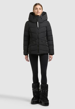 Femme portant une veste d'hiver matelassée noire avec capuche, des leggings noirs et des bottes de neige isolées noires, debout sur un fond gris clair.