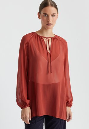 Blouse rouge transparente à manches longues et poignets élastiques. Dotée d'une fermeture à nouer au cou. Tissu doux et léger avec une coupe ample.