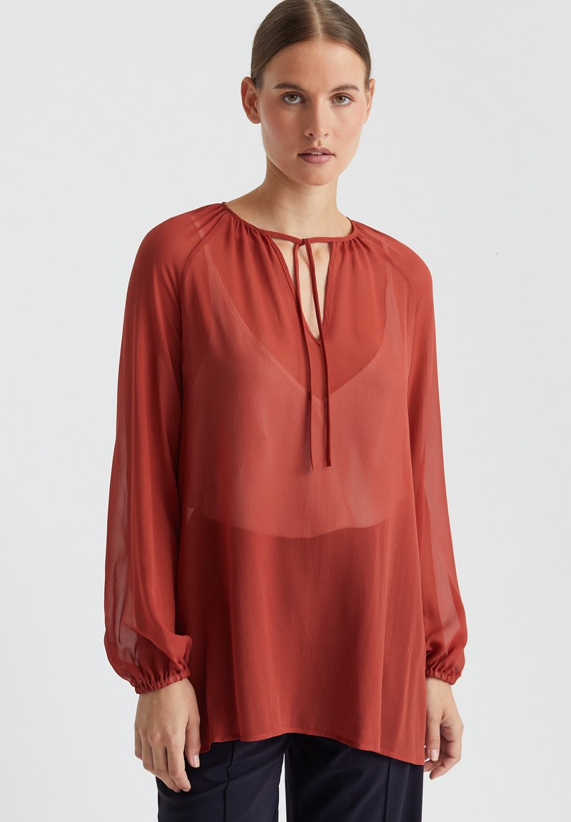 Rote transparente Bluse mit langen Ärmeln und elastischen Bündchen. Mit einer Schleifen-Nackenverschluss versehen. Aus weichem, leichtem Stoff mit lockerer Passform.
