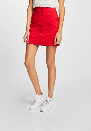 Femme portant une mini-jupe rouge boutonnée sur le devant, associée à des baskets blanches et un haut blanc rentré, debout devant un fond uni.