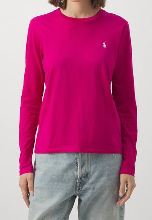 Woman wearing a bright pink long-sleeve shirt with a small white embroidered logo on the left chest, paired with light blue jeans.
