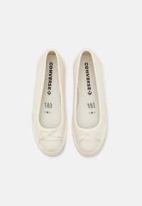Scarpe slip-on in tela bianca con punta rotonda, sottile dettaglio con fiocco sulla parte superiore e marchio testuale sulla soletta. Superficie liscia, design minimalista.