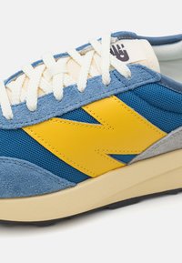 Blå sneakers med gula accenter, mesh- och mockamaterial, krämfärgade snören, och en beige gummisul. Har en upphöjd logga på tungan.