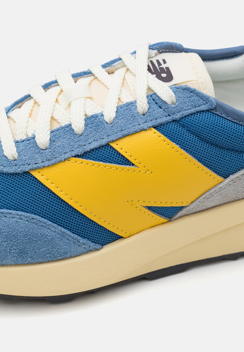 Zapatilla azul con acentos amarillos, materiales de malla y ante, cordones color crema y suela de goma beige. Presenta un logo en relieve en la lengüeta.