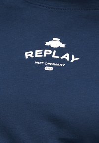 Ύφασμα σε ναυτικό μπλε με λευκό τυπωμένο κείμενο: «REPLAY», «NOT ORDINARY» και «1981» κάτω από ένα γραφικό με στυλιζαρισμένη κορδέλα και έμβλημα.