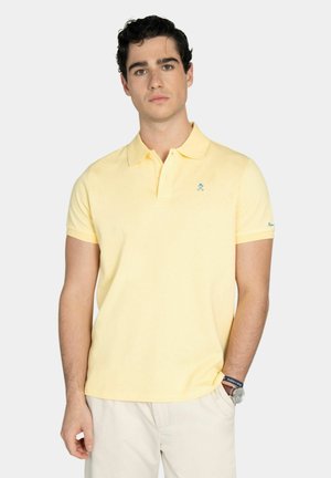 Joven con camisa polo de manga corta color amarillo pálido y pantalones beige claro, de pie con una mano en el bolsillo, fondo neutro.
