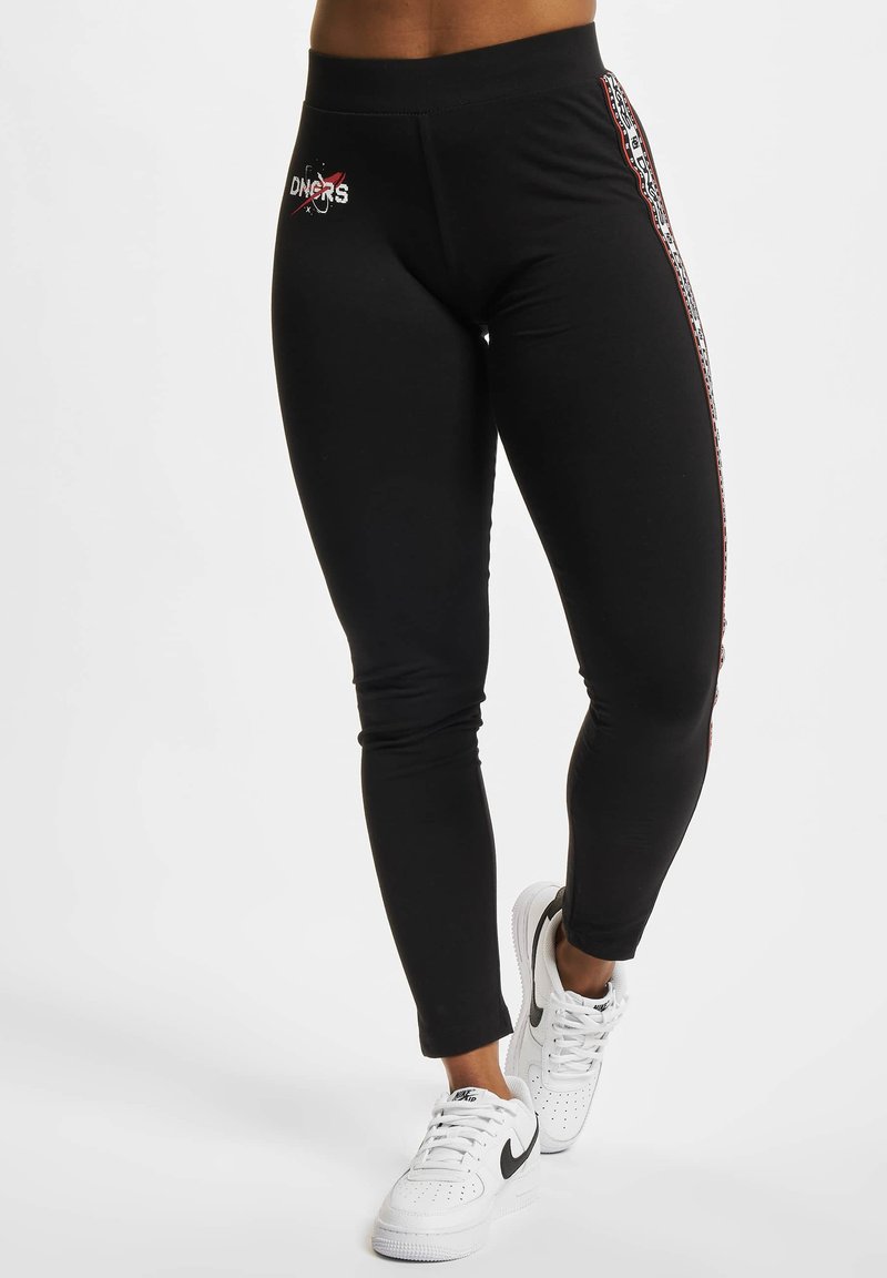 Svart leggings med en tettsittende design, med et brodert logo foran og et mønstret sidestripe. Nervest med hvite sneakers.