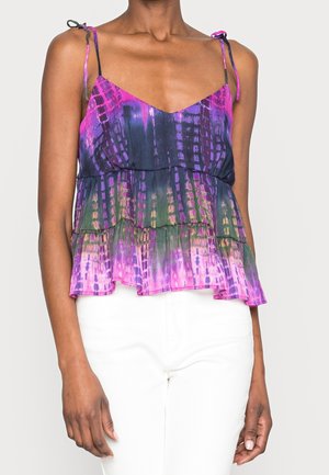 Nainen, jolla on yllään hihaton, solmittavilla olkapäillä varustettu paita, jossa on violetteja, pinkkejä, vihreitä ja mustia batikkikuvioita, yhdistettynä valkoisiin housuihin, seisoo yksinkertaisen taustan edessä.