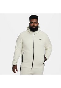 Bluza Nike z kapturem w kolorze ecru, z zamek błyskawicznym z przodu, czarnymi akcentami na kieszeniach oraz miękkim, teksturowanym materiałem.