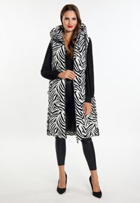 Zebra-mønstret puffer vest med hætte, sort og hvidt stof, stående længde, lynlås lukning, parret med tætsiddende sorte bukser og højhælede sko.