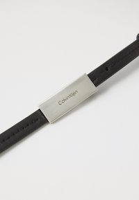Bracciale in pelle nera con una piastra rettangolare in metallo argento incisa con "Calvin Klein". Texture liscia e chiusura sicura.