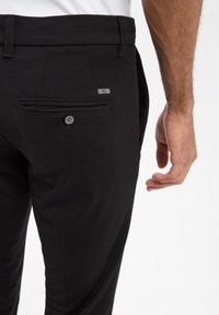 Pantalons noirs en tissu texturé, dotés d'une poche arrière avec un bouton et d'une petite étiquette en métal.