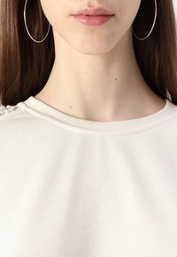 Gaišas krēma sporta krekls ar ribotu dekoltē un dekoratīvām pērļu akcentiem vienā plecā. Gluda auduma, relaksēta piegriezuma.