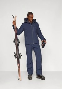 Άνδρας ντυμένος με ναυτικό μπλε σκι outfit κρατώντας σκι με το ένα χέρι και γυαλιά σκι με το άλλο, φορώντας μαύρες μπότες σκι, στέκεται σε άσπρο πάτωμα.