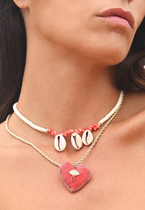 Deux colliers superposés : l'un est orné de coquillages blancs et de perles corail, l'autre d'un pendentif en forme de cœur rouge avec des accents décoratifs sur une chaîne en or.