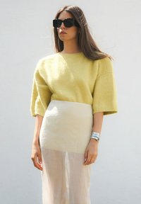 Gelber Strickpullover mit kurzen, weiten Ärmeln, kombiniert mit einem cremefarbenen, fließenden Rock. Das Model trägt große schwarze Sonnenbrillen und silberne Armbänder.