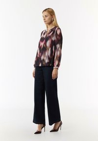 Langärmlige Bluse mit einem mehrfarbigen abstrakten Druck, plissiertem Design und Schlüssel-Ausschnitt, kombiniert mit weiten, dunklen Jeans und spitzen Absätzen.