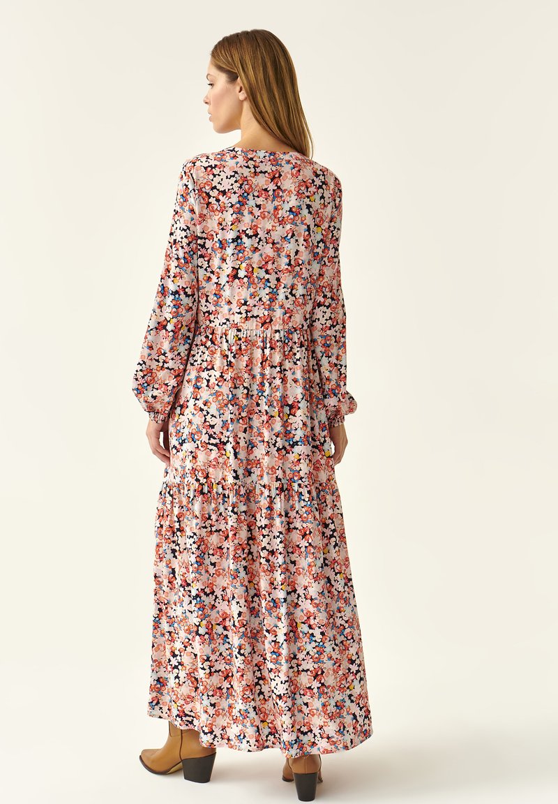 Maxikleid mit langen Ärmeln, das ein farbenfrohes Blumenmuster auf einem hellrosa Hintergrund und einen gestuften Saum aufweist. Getragen mit braunen Knöchelstiefeln.