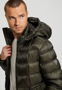 Puffer jacket in dark green, featuring horizontal quilted sections, a detachable hood, and zip closure. Includes a patch logo on the sleeve.