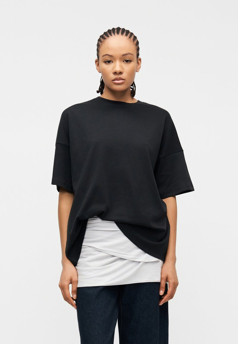 Νεαρό άτομο με πλεξούδες, φοράει φαρδιά μαύρη μπλούζα πάνω από ένα στρωματοποιημένο λευκό τοπ και σκούρο παντελόνι, κοιτάζοντας μπροστά σε απλό φόντο.