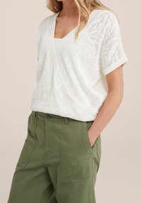 Top en crochet blanc avec un col en V et des manches courtes, présentant un motif délicat et une coupe ample. Associé à un pantalon cargo vert.