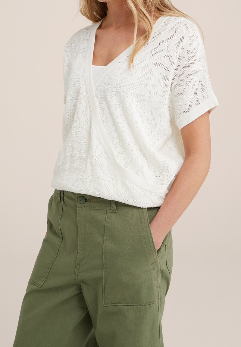 Top en crochet blanc avec un col en V et des manches courtes, présentant un motif délicat et une coupe ample. Associé à un pantalon cargo vert.