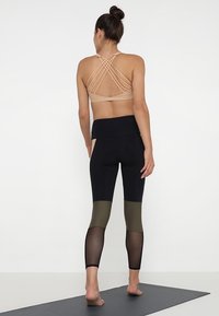 Jasno beżowy sportowy stanik na ramiączkach z krzyżującym się tyłem, zestawiony z czarnymi legginsami z oliwkowymi akcentami i przezroczystymi panelami z siateczki.