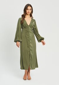 Olivno zeleni satenski čim z globokim V-izrezom, dolgimi napihnjenimi rokavi, sprednjimi gumbi in razporkom. Tkanina ima gladko, sijoče teksturo.