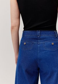 Niebieskie dżinsy o wysokim stanie z dwoma tylnymi kieszeniami, z metalowymi guzikiem i widocznymi detalami szwów. Gładka faktura, dopasowany krój.