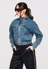 Femme avec queue de cheval portant une veste en jean oversize, de grandes boucles d'oreilles géométriques dorées, un pantalon noir et de grandes lunettes de soleil noires, les mains dans les poches.