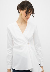 Blusa avvolgente bianca con maniche lunghe, scollatura a V profonda e dettaglio con bottone laterale. Realizzata in tessuto morbido con un orlo asimmetrico drappeggiato.