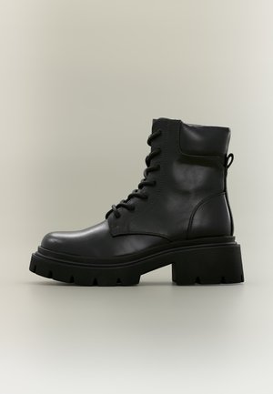 Botte de combat noire en cuir à lacets avec semelle épaisse en caoutchouc et talon bloc surélevé, vue latérale sur un fond neutre uni.