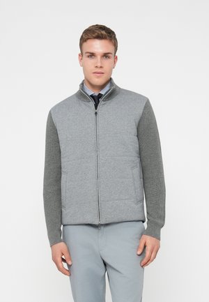 Chaleco gris con cremallera y parte delantera acolchada, con mangas de canalé texturizadas. Cuenta con dos bolsillos laterales y un cuello alzado, llevado sobre una camisa de cuadros.