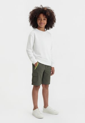 Göndör hajú gyermek fehér pulóvert, olívazöld cargo rövidnadrágot zöld cipzárdísszel és fehér sportcipőt viselve, egyik kezével a zsebében áll.