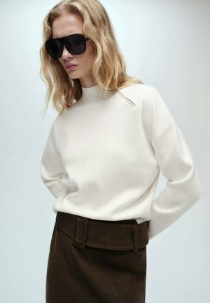 Cream sweater with a high neck, paired with a dark brown skirt featuring a textured wide belt. Model wears oversized black sunglasses.