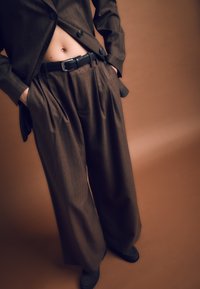 Hnedý pruhovaný oblek na mieru so širokými nohavicami a priliehavým blejzrom, doplnený o čierny opasok a hladkú textúru látky.