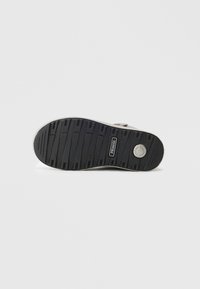 Semelle de chaussure en caoutchouc noir avec des rainures horizontales et un logo circulaire. Bord extérieur lisse avec une zone de talon renforcée.