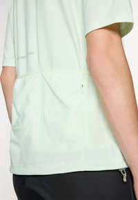 Camisola atlética verde claro com mangas curtas, tecido texturizado, bolso traseiro elástico e bainha reta. Apresenta um pequeno detalhe com zíper.