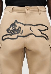 Μπεζ παντελόνια εργασίας από ανθεκτικό ύφασμα με μεγάλο μαύρο γκραφίτι σπρέι που απεικονίζει ένα ζώο που πηδάει στην πίσω πλευρά. Δύο εμφανείς μπροστινές τσέπες.