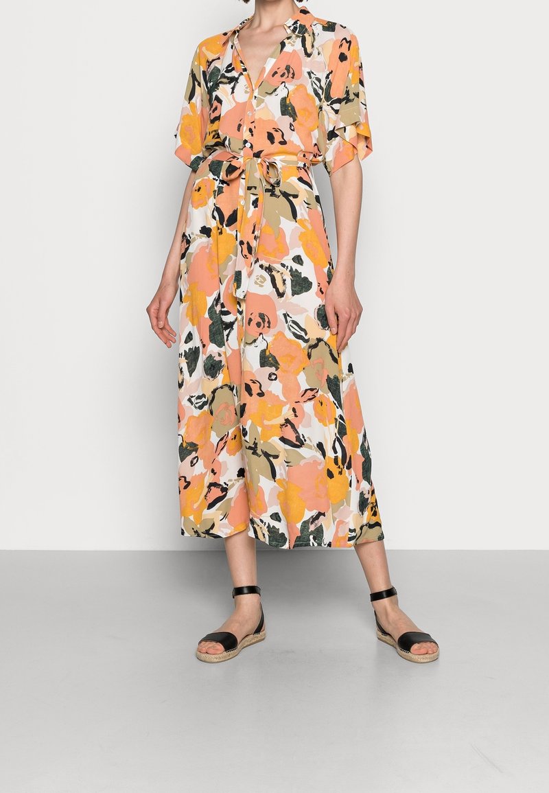 Femme portant une robe midi fleurie aux tons orange, rose et vert, avec une ceinture à la taille, des manches courtes et des sandales noires à bride cheville.
