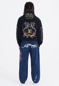 Svart huvtröja med färgglad drakbroderi och text på baksidan; blå denimbyxor med vit logotyp och siddekoration; avslappnad passform.