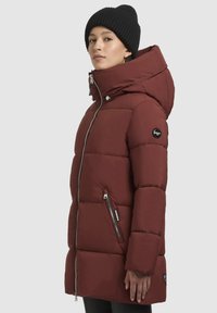 Rød isoleret hættejakke med quiltet design, frontlynlås, sidelommer og et sort logo-patch på ærmet. Sort strikket hue båret.